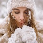Close-up of a woman in winter attire surrounded by snow, blowing warm air into her hands.