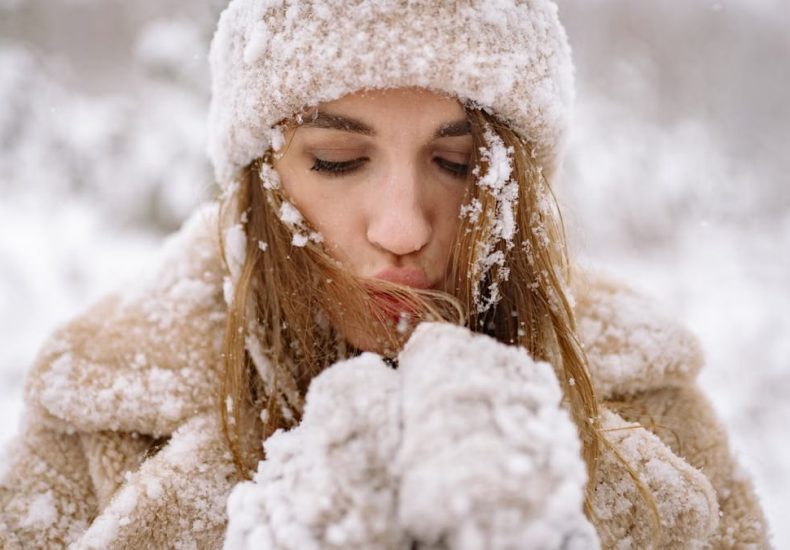Close-up of a woman in winter attire surrounded by snow, blowing warm air into her hands.